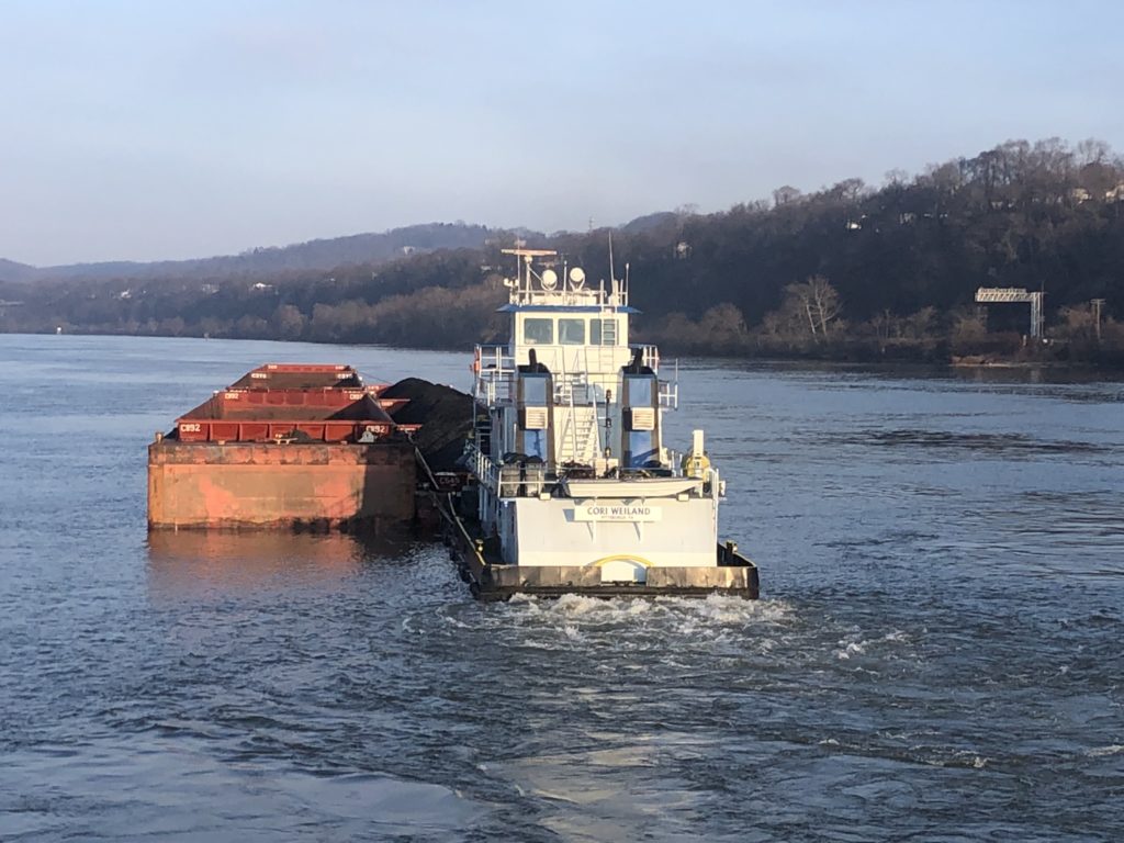 The towboat Cori Weiland pushing four barges of coal and stone up a wide river at low sun, hills on the far bank. CORI WEILAND is lettered on the stern.