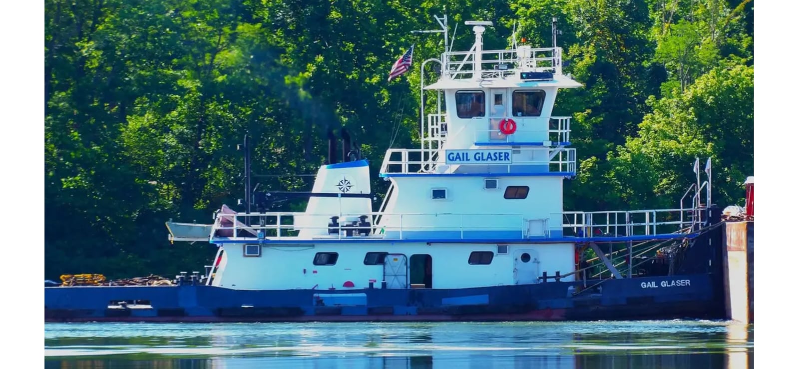 Gail Glaser alongside a barge, its white superstructure and blue hull reflected in the river below a wooded bank.
